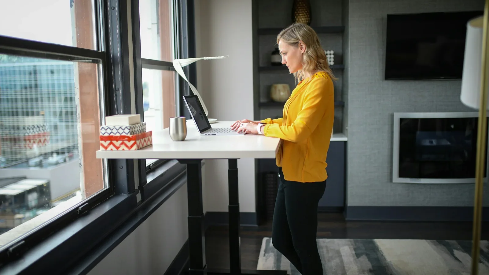 A woman working on a laptop at a standing desk beside an office window